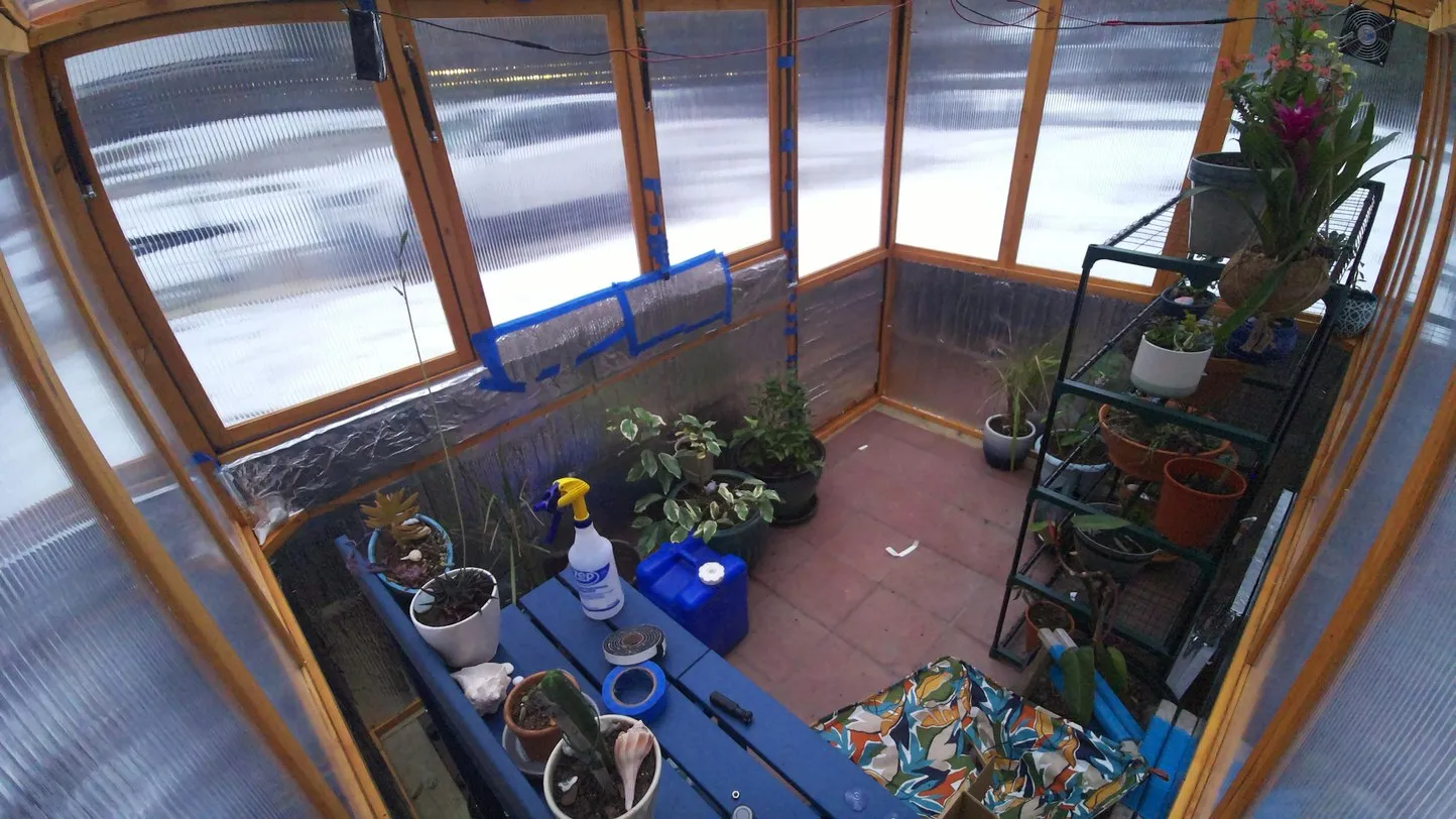 Inside the greenhouse: a wood-framed polycarbonate structure with shelves of potted plants, a blue workbench with tools and shell-filled pots, hanging baskets, a small circulation fan, and sensor wiring taped along the frame. The sound glows through the translucent walls.